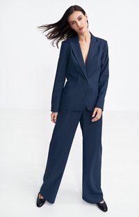 Granatowy garnitur w prążki z dopasowaną marynarką i szerokimi spodniami, z pojedynczym zapięciem na guzik i eleganckimi czarnymi butami.