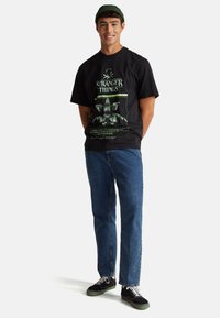 Černé tričko s grafickým motivem "Stranger Things" v zelené barvě. Pohodlný střih, kombinované s modrými džínami a černými teniskami s bílými tkaničkami.