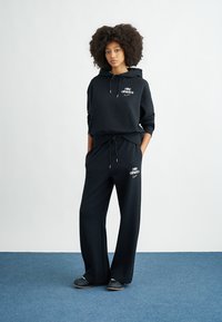 Μαύρο φούτερ με κουκούλα και τσέπη, σε συνδυασμό με μαύρο φαρδύ παντελόνι φόρμας με λευκά λογότυπα και στα δύο. Λείο ύφασμα, άνετη εφαρμογή.