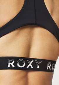 Černá sportovní podprsenka se širokým elastickým páskem s logem "ROXY" v bílé barvě. Hladká tkanina, minimalistický design a tvar s racerback.