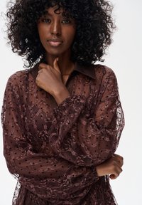 Burgundowa, przezroczysta bluzka z koronkowym wykończeniem, w kropki, z delikatnie falbankowymi mankietami; ma kołnierzyk i lekką teksturę.