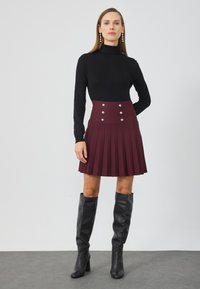 Sort rullekravebluse parret med en maroon plisseret nederdel, der har fem sølvknapper i taljen. Modellen har knæhøje sorte støvler på.