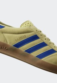 Žluté semišové tenisky s modrými koženými pruhy, mají texturovaný jazyk a hnědou gumovou podrážku, označené "GAZELLE" na boku.