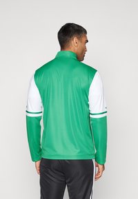 Groene sportjas met een hoge kraag, lange witte mouwen en groene gestreepte accenten op de mouwen, gemaakt van een gladde, lichte stof.
