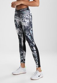 Piernas conleggings Nike Pro de patrones abstractos en blanco y negro y zapatillas Nike blancas con acentos en verde neón sobre un fondo plano.