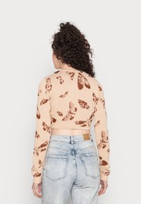 Sweater curto em bege claro com padrões de borboletas castanhas; textura de malha, mangas longas e punhos canelados, combinado com jeans de cintura alta em azul claro.