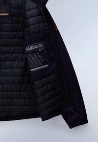 Chaqueta de plumas negra con textura acolchada en la mitad superior, mitad inferior suave y cierre de cremallera. Presenta etiquetas internas y acentos brillantes.