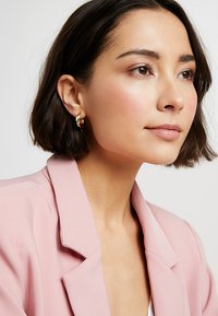 Boucles d'oreilles en or en forme de cercle et blazer rose avec revers. Texture lisse et design contemporain, mettant en valeur les contours doux et l'éclat subtil.