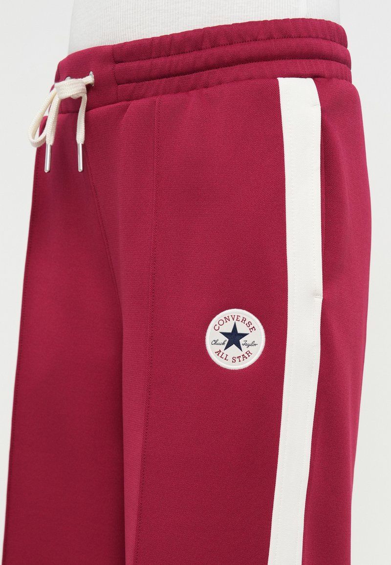 Vinrøde joggingbukser med hvid stribe, elastisk talje og snor. Har et rundt Converse-logo på låret.