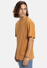 Jonge man met schouderlang krullend haar, gekleed in een bruine korte mouw t-shirt en blauwe broek, kijkend naar links.