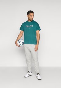 Camiseta de algodão azul-água com estampa gráfica, calças de moletom cinzas, tênis preto e branco, segurando uma bola de futebol branca com detalhes azuis.
