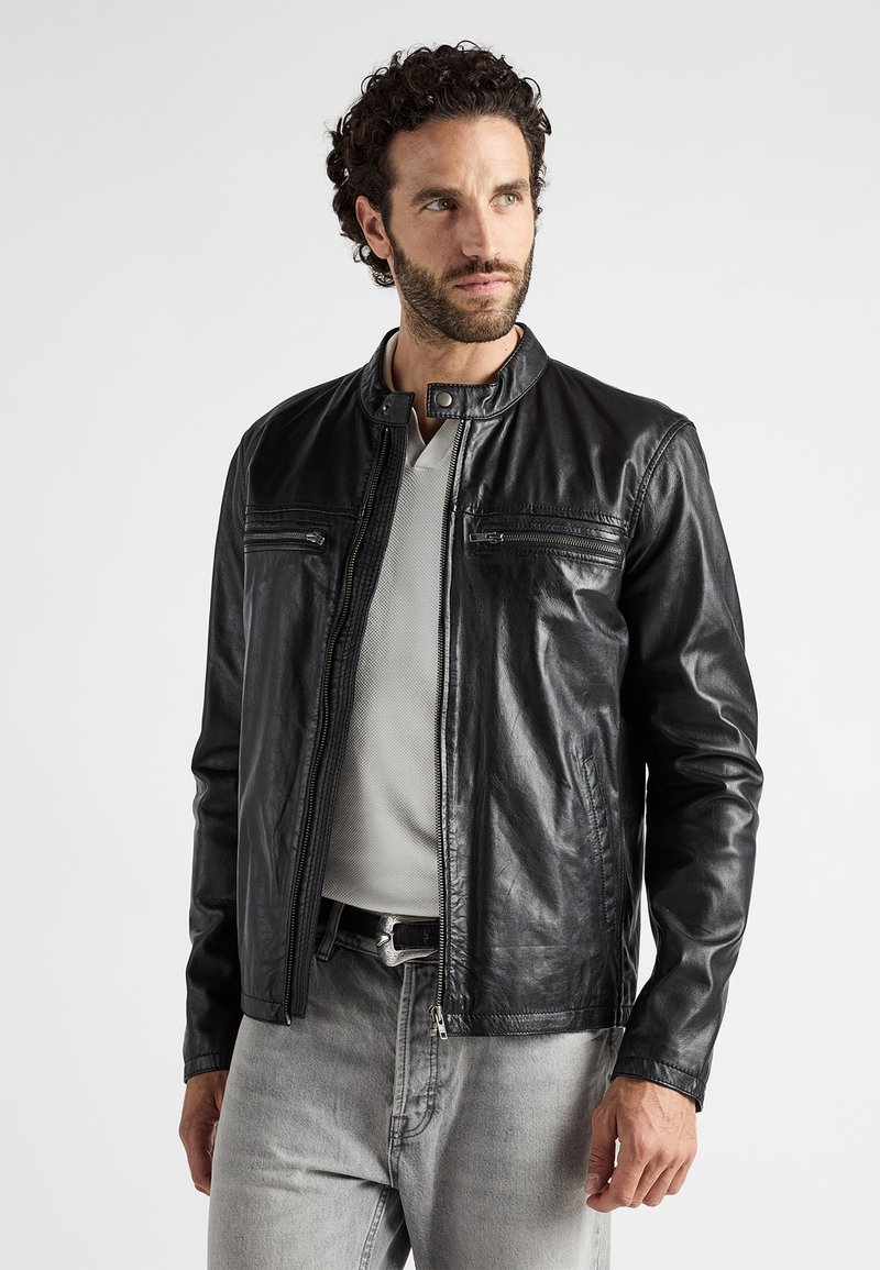 Schwarze Lederjacke mit Stehkragen, zwei verschließbaren Brusttaschen und langen Ärmeln. Getragen über einem hellgrauen Hemd und Jeans.