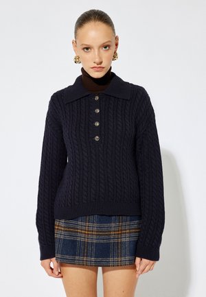Granatowy sweter w splot warkoczowy z kołnierzykiem, pięcioma guzikami i ściągaczami u rękawów, w połączeniu z mini spódnicą w kratę w granatowo-brązowych odcieniach.