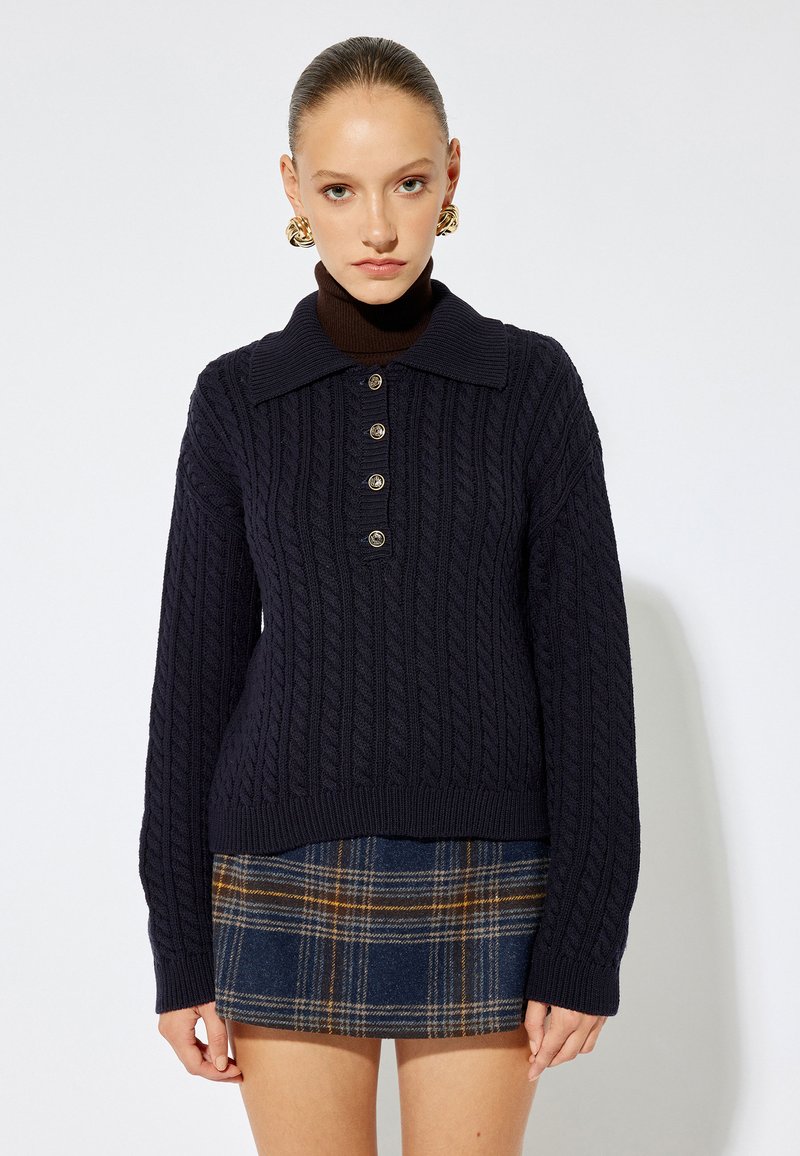 Marineblaue Kabelstrickpullover mit Kragen, fünf Knöpfen und gerippten Bündchen, kombiniert mit einem karierten Minirock in Marineblau- und Brauntönen.