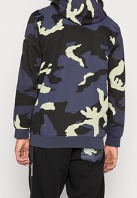 Baksidan av en person som bär en blå, svart och beige camouflage hoodie och svarta byxor med kamouflagefickdetalj.