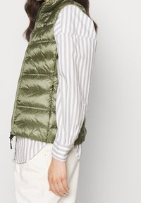 Pikowana kamizelka w oliwkowej zieleni z wysokim kołnierzem. Strukturystyczny, pikowany wzór. Noszona na jasnej, pasiastiej koszuli z długimi rękawami. Zapinana na suwak po bokach.