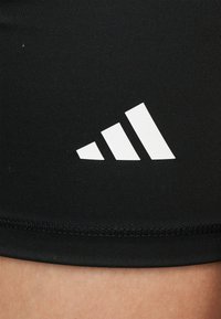 Logo Adidas blanc à trois bandes sur un tissu de sport côtelé noir au-dessus de la peau nue d'une personne.