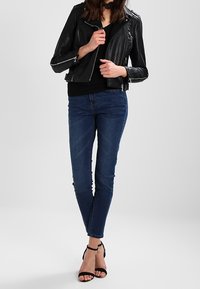 Giacca in pelle nera da motociclista con tasche con zip, abbinata a jeans slim-fit blu scuro. Tacchi alti con cinturino alla caviglia neri, design minimalista.