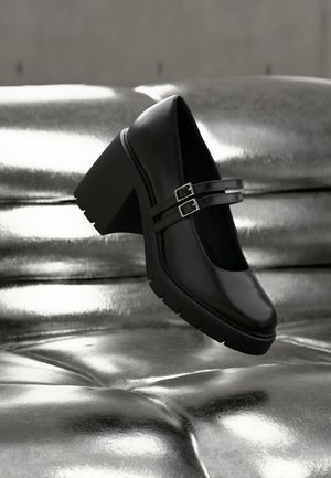 Chaussure noire à talon épais avec double bride à boucle reposant sur une surface argentée capitonnée brillante, mettant en valeur la texture et le design de la chaussure.