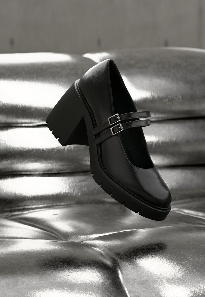 Μαύρο παπούτσι με χοντρό τακούνι και διπλές λουρίδες με αγκράφες, τοποθετημένο σε γυαλιστερή ασημένια επιφάνεια με κουμπιά, που αναδεικνύει την υφή και το σχέδιο του παπουτσιού.