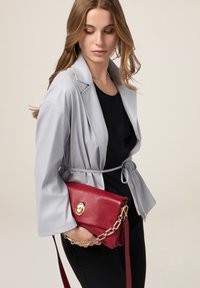 Giacca grigia chiaro con cintura e maniche larghe, abbinata a un top nero e a una borsa rossa strutturata con hardware dorato e tracolla a catena.