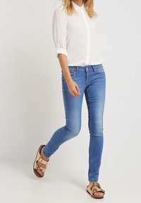 Persona vestida con una camisa blanca de manga larga, jeans skinny azules y sandalias metálicas de color rosa dorado caminando frente a un fondo claro y liso.
