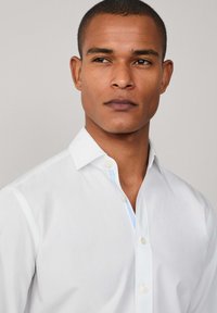 Camisa blanca de botones con una textura suave, que presenta un cuello clásico y sutiles acentos azules en el interior de la tapeta.