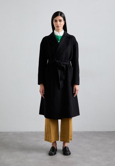 Abrigo largo negro con cintura ajustada, bolsillos laterales y cuello de solapa. Llevado sobre un suéter verde y pantalones de pana mostaza, combinado con mocasines negros.