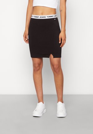 Kvinde iført sort nederdel med Tommy Jeans mærket taljebånd, hvid crop top og hvide sneakers, stående på gråt gulv foran hvid væg.