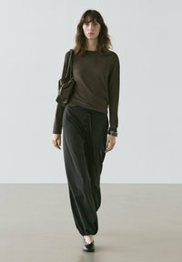 Kvinde går fremad iført mørkebrun langærmet top, løse sorte bukser med knapper, sorte sko, bærende en brun skuldertaske med tekstur.