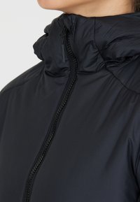Veste rembourrée noire avec un col haut, fermeture éclair, et un tissu lisse et léger avec un léger reflet.