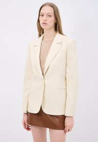 Blazer crema con rever a punta, due tasche frontali e silhouette snella. Indossato sopra un top beige e una gonna in pelle marrone.