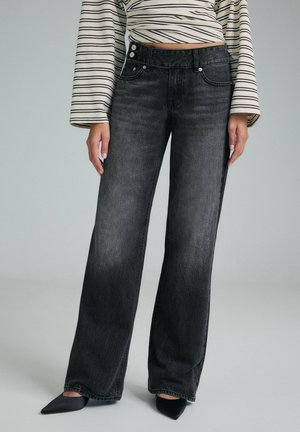Femme portant un jean noir taille haute à jambes larges, des chaussures noires pointues et un crop top rayé à manches longues, debout devant un fond uni.