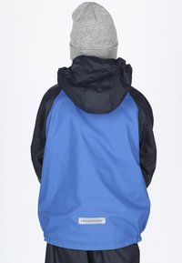 Person iført en blå og sort hættetrøje og en grå strikket hue, vendt væk på en ensfarvet hvid baggrund.