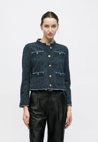 Giacca corta blu testurizzata con collo tondo, bottoni dorati e due tasche frontali. Presenta bordi sfrangiati e maniche lunghe.