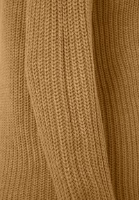 Maglione lavorato a maglia in un caldo colore cammello con una texture a coste e cuciture visibili. Il tessuto appare morbido e spesso.