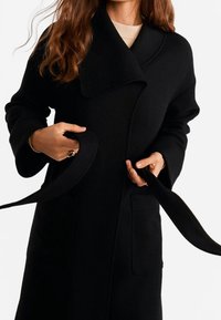 Černý vlněný kabát s velkým límcem, dvěma předními kapsami a opaskem. Textilie vypadá silně a strukturovaně.