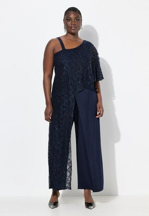 Navy jumpsuit featuring one shoulder design, sheer lace overlay with sequins, wide legs, and a textured finish. Smooth fabric on the leg.