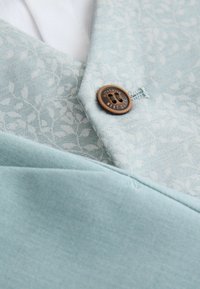 Tissu bleu clair avec un motif subtil de feuilles et un bouton marron portant la mention « Authentic Next Duke » cousu près d'une boutonnière.