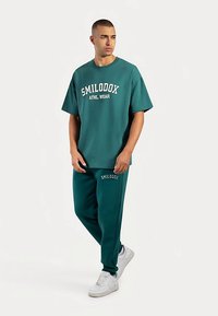 Ensemble de vêtements de sport bleu sarcelle comprenant un t-shirt à manches courtes avec un logo blanc et un pantalon de jogging assorti, tous deux en tissu doux avec une coupe décontractée.
