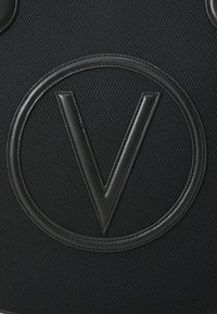 Borsa in tessuto nero con un grande logo circolare in rilievo con la lettera "V" al centro, realizzata in materiale testurizzato con dettagli in pelle.