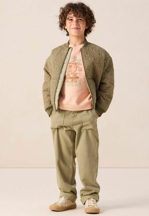 Olivgrüne, gesteppten Bomberjacke über einem pfirsichfarbenen Sweatshirt mit Grafik, kombiniert mit hellgrünen Cargo-Hosen und beigen Sneakern.