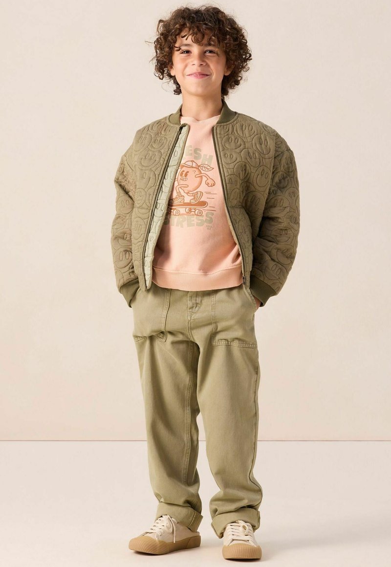Blouson aviateur matelassé vert olive, sweat-shirt graphique rose pâle avec un dessin animé, pantalon cargo beige assorti, et baskets beiges.