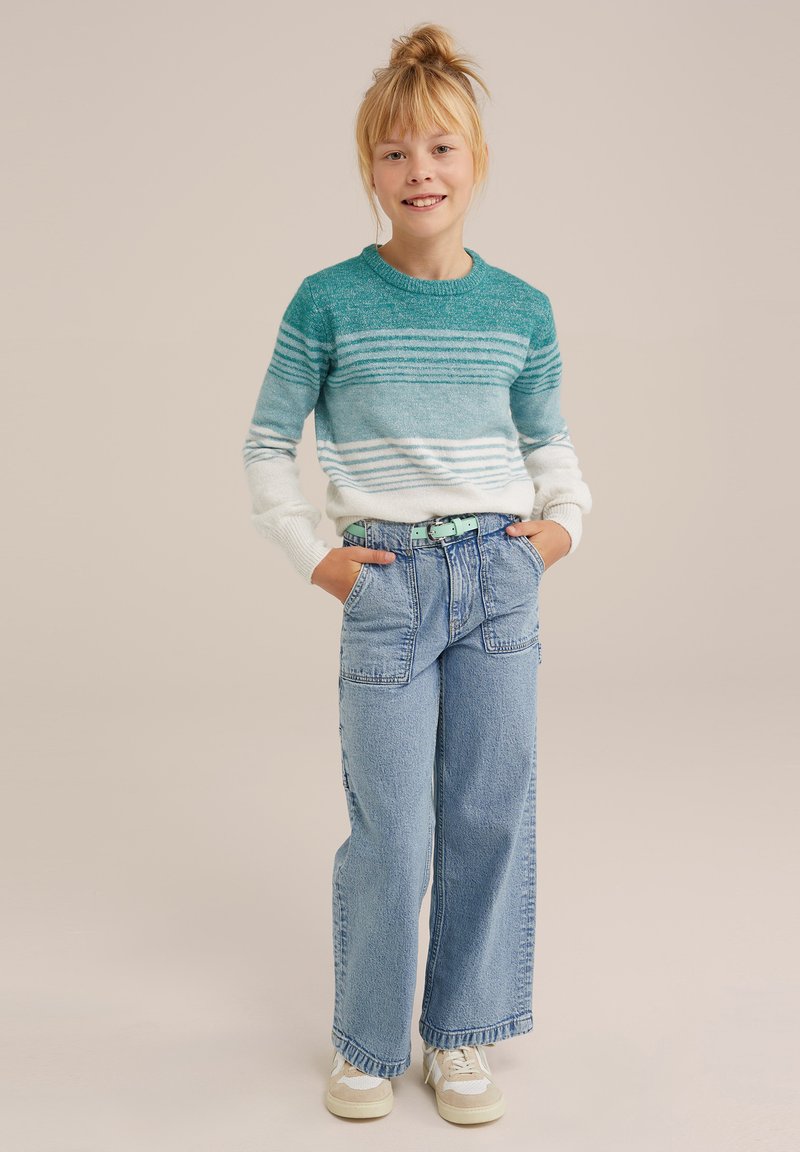 Hellblauer und weiß gestreifter Pullover, lockere blaue Jeans mit Taschen und weiße Sneakers mit cremefarbigen Akzenten. Mädchen steht mit Händen in den Taschen.