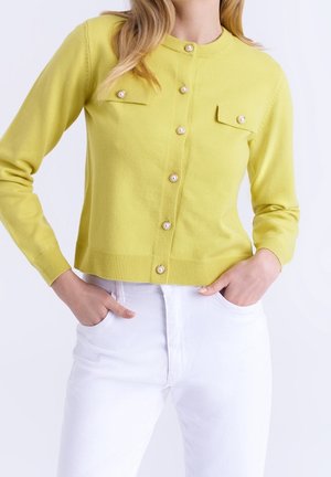 Žena v limetkově zeleném kardiganu na knoflíky s perlovými knoflíky a kapsami s klopami, v kombinaci s bílými kalhotami s vysokým pasem, ruce v kapsách.