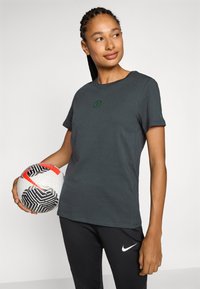 Mörkgrå t-shirt i mjukt tyg med rund hals, korta ärmar och en grön logotyp på bröstet. Modellen håller en mönstrad fotboll.