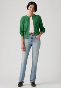 Πράσινη ζακέτα με πλέξη καλώδιο, στρογγυλή λαιμόκοψη, κλείσιμο με κουμπιά και μανσέτες με ριμπ ύφανση, σε συνδυασμό με τζιν παντελόνι ελαφρού μπλε, ίσιας γραμμής και καφέ παπούτσια.