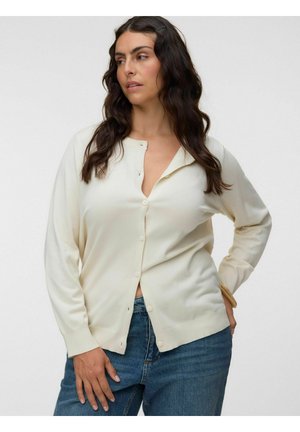 Frau mit langen dunklen Haaren, die eine elfenbeinfarbene, geknöpfte Strickjacke und blaue Jeans trägt, posiert mit einer Hand auf der Hüfte und schaut seitlich.