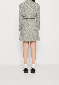 Vestido de estampado floral en verde apagado; mangas largas, cintura ajustada, falda acampanada. Combinado con calcetas blancas y zapatos negros gruesos.