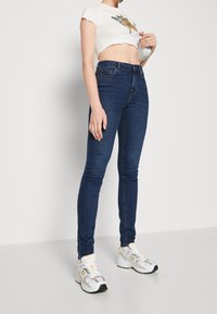 Úzké modré džíny s vysokým pasem, doplněné krátkým bílým tričkem s grafickým motivem a bílými teniskami se šedými detaily.